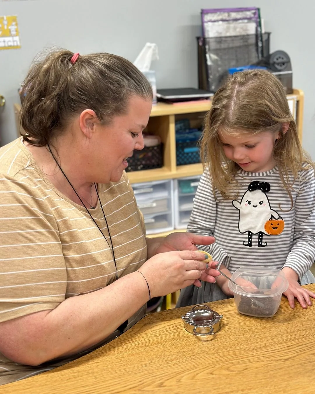 An adult woman with long brown hair in a ponytail and a young girl with blonde hair are working together at a table. The woman is wearing a tan striped shirt and the girl is wearing a striped shirt with a ghost and pumpkin design. They seem to be engaged in a crafting or baking activity, with a small metal cookie cutter and a plastic container on the table. The background shows shelves with storage bins and a few office supplies.