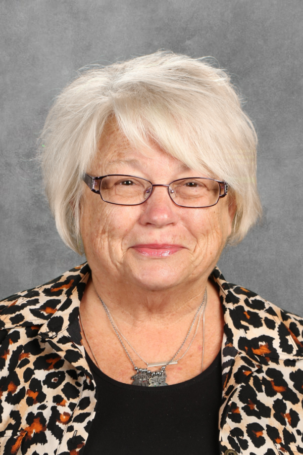 An elderly woman with short white hair, glasses, and a leopard print shirt, smiling at the camera against a gray background.