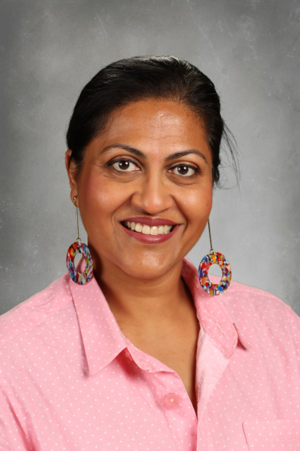 A woman with dark hair pulled back, wearing a pink polka dot shirt and colorful hoop earrings, smiling against a gray background.