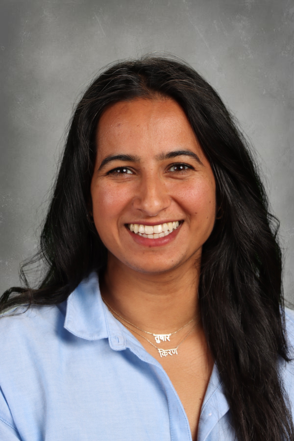 A woman with long black hair, wearing a light blue collared shirt and layered necklaces with Hindi words, smiling against a gray background.