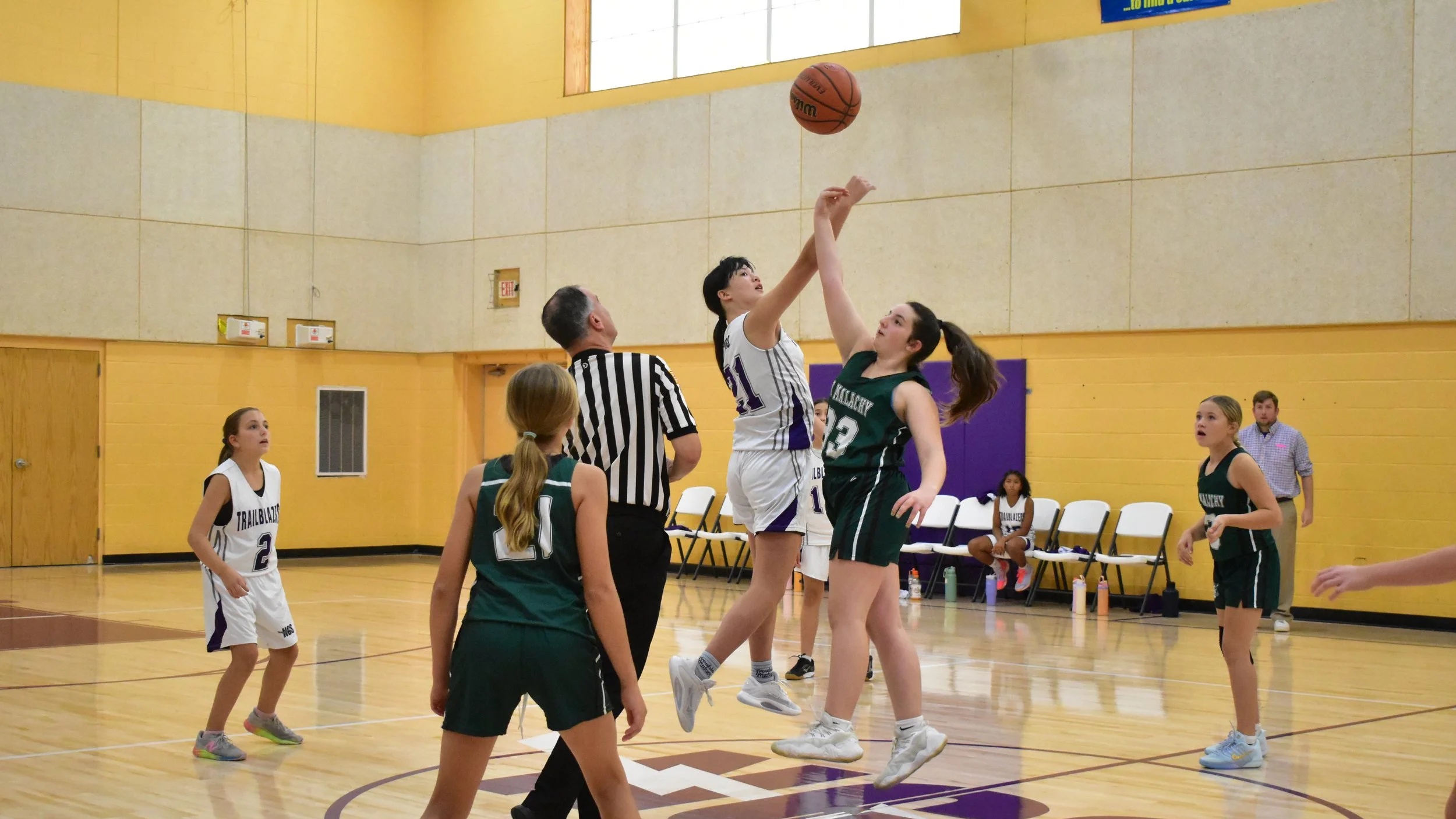Young girls playing basketball in an indoor gym, with two players jumping for the ball at the tip-off, officials, and spectators.