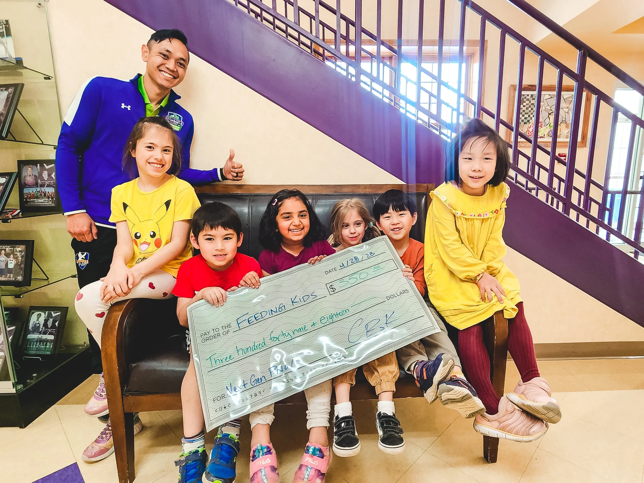 Six children sitting and standing on a bench, holding a large check for $3500, with an adult man standing behind them giving a thumbs-up. The children are smiling and appear happy, in a room with photos on a shelf and a staircase in the background.