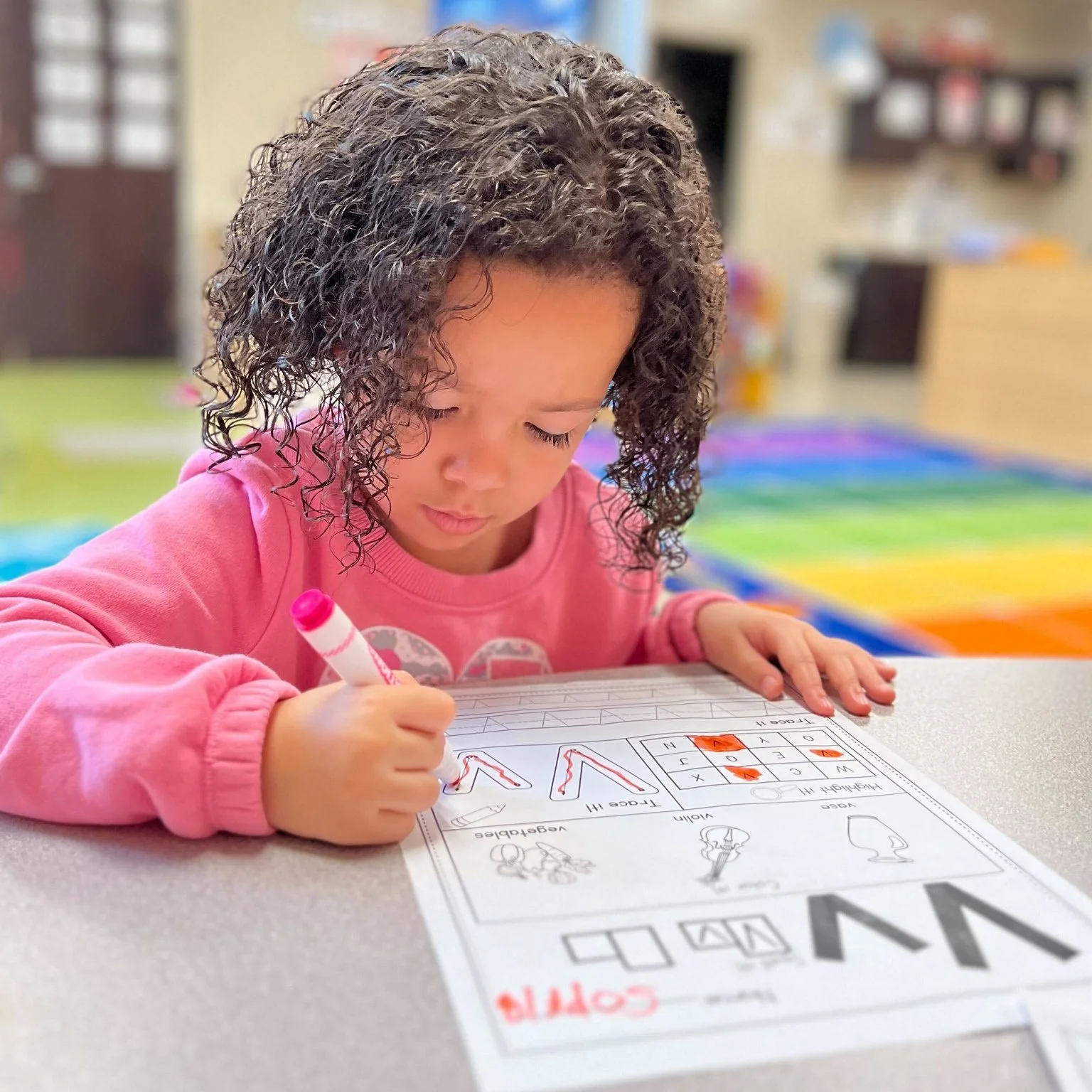 A young girl with curly hair and a pink sweatshirt is coloring a worksheet with a pink marker at a table in a classroom.
