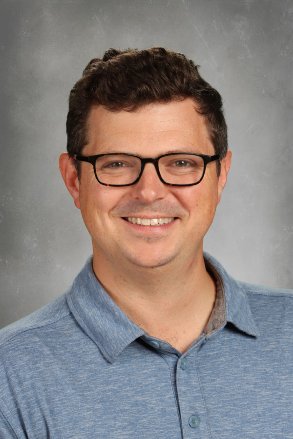 A smiling man with short dark hair, wearing black glasses and a blue collared shirt, against a gray background.