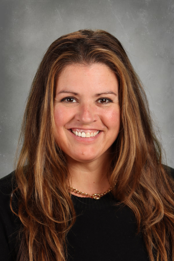 Portrait of a woman with shoulder-length wavy brown hair, smiling, wearing a black top and gold chain necklace, against a gray background.