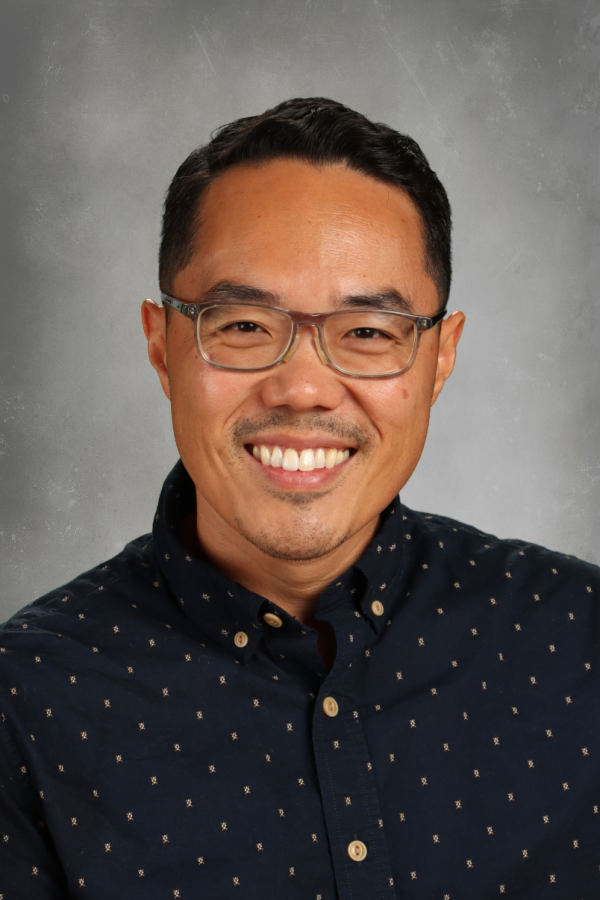 A smiling Asian man wearing glasses and a navy blue button-up shirt with small white patterns, posing against a gray background.