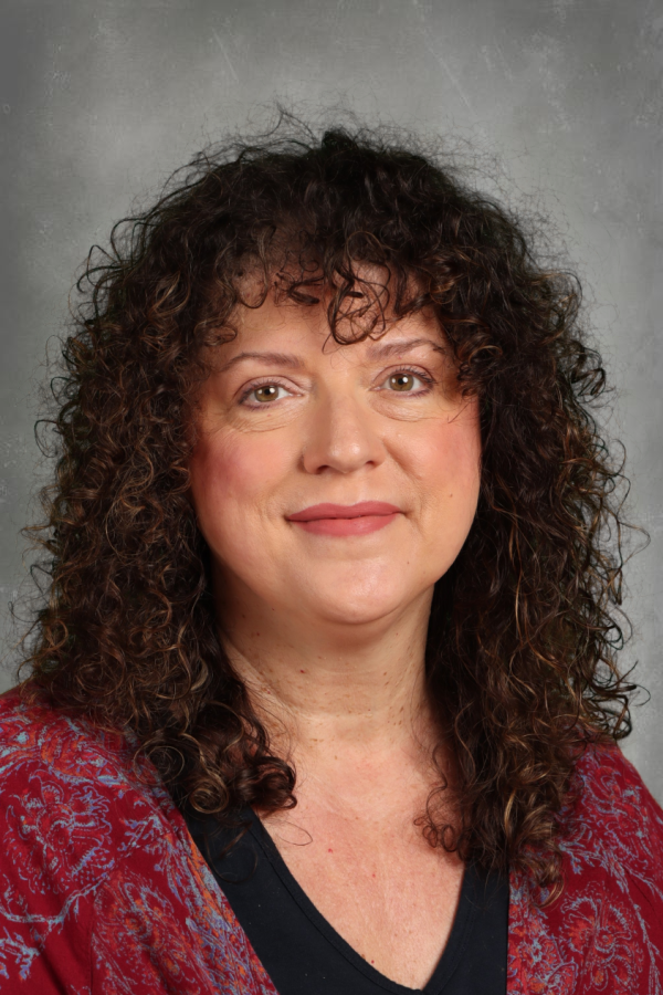 A woman with curly brown hair and light skin, wearing a red patterned jacket and black top, smiling at the camera against a gray background.