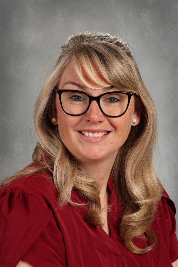 A woman with blonde hair, glasses, and pearl earrings smiling in a red shirt against a gray background.