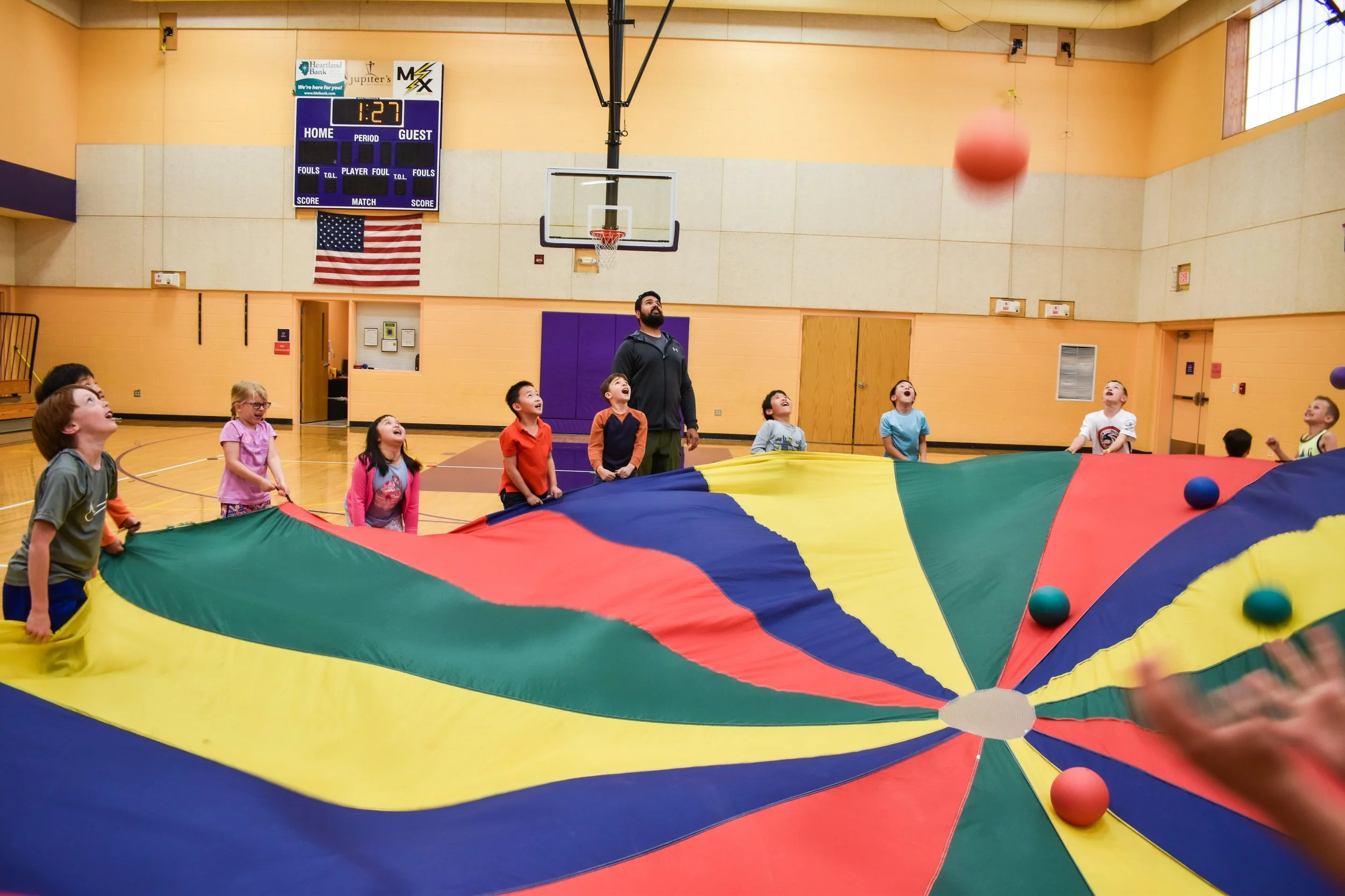 Children and an adult playing with a colorful parachute inside a gymnasium, with balls on the parachute, basketball hoop, scoreboard, and American flag visible in the background.