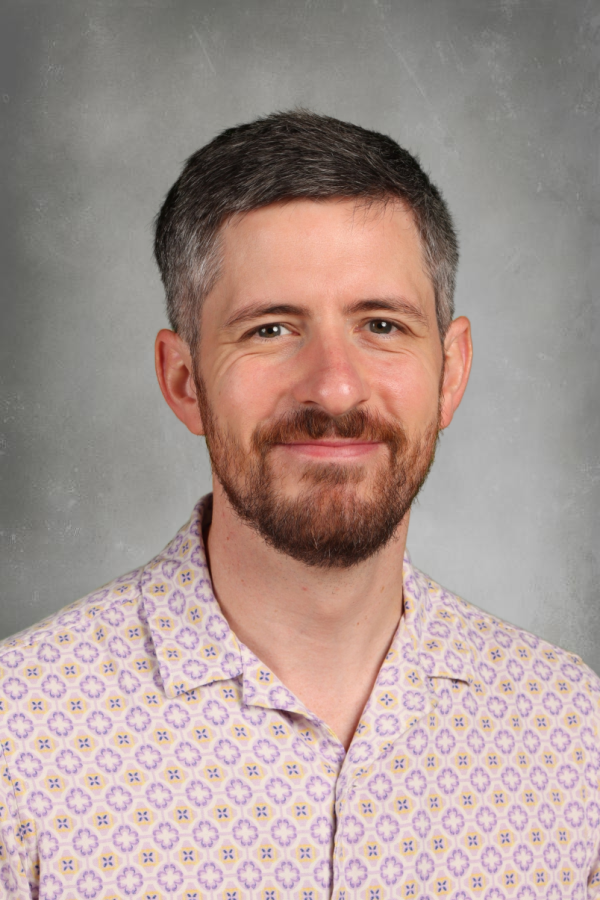 Portrait of a man with short brown hair and a beard, smiling, wearing a patterned shirt with purple, yellow, and white colors, against a plain gray background.