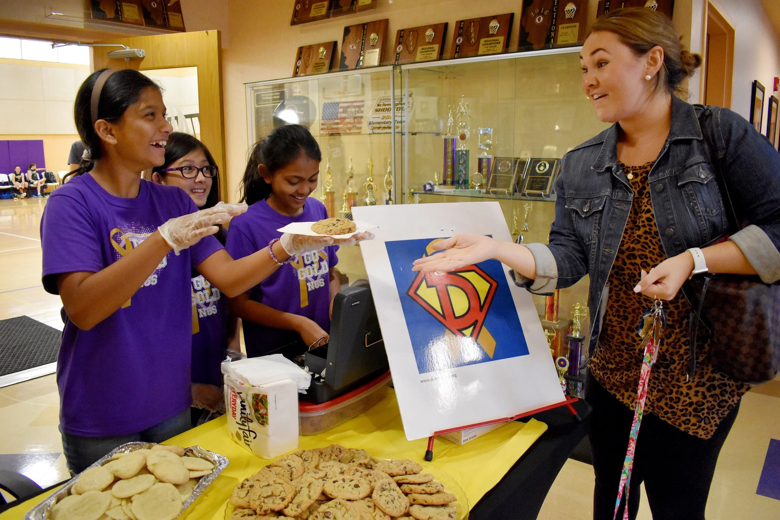 Girls in purple shirts selling cookies at a fundraiser table, with an adult woman in a denim jacket talking to them. There are cookies on the table and a sign with a superman logo.
