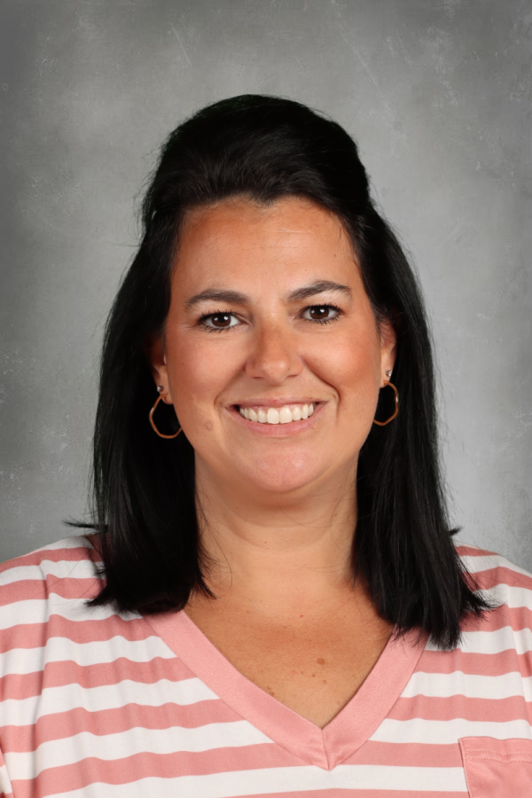 A woman with dark hair, wearing gold hoop earrings and a pink and white striped V-neck shirt, smiling at the camera against a plain, gray background.