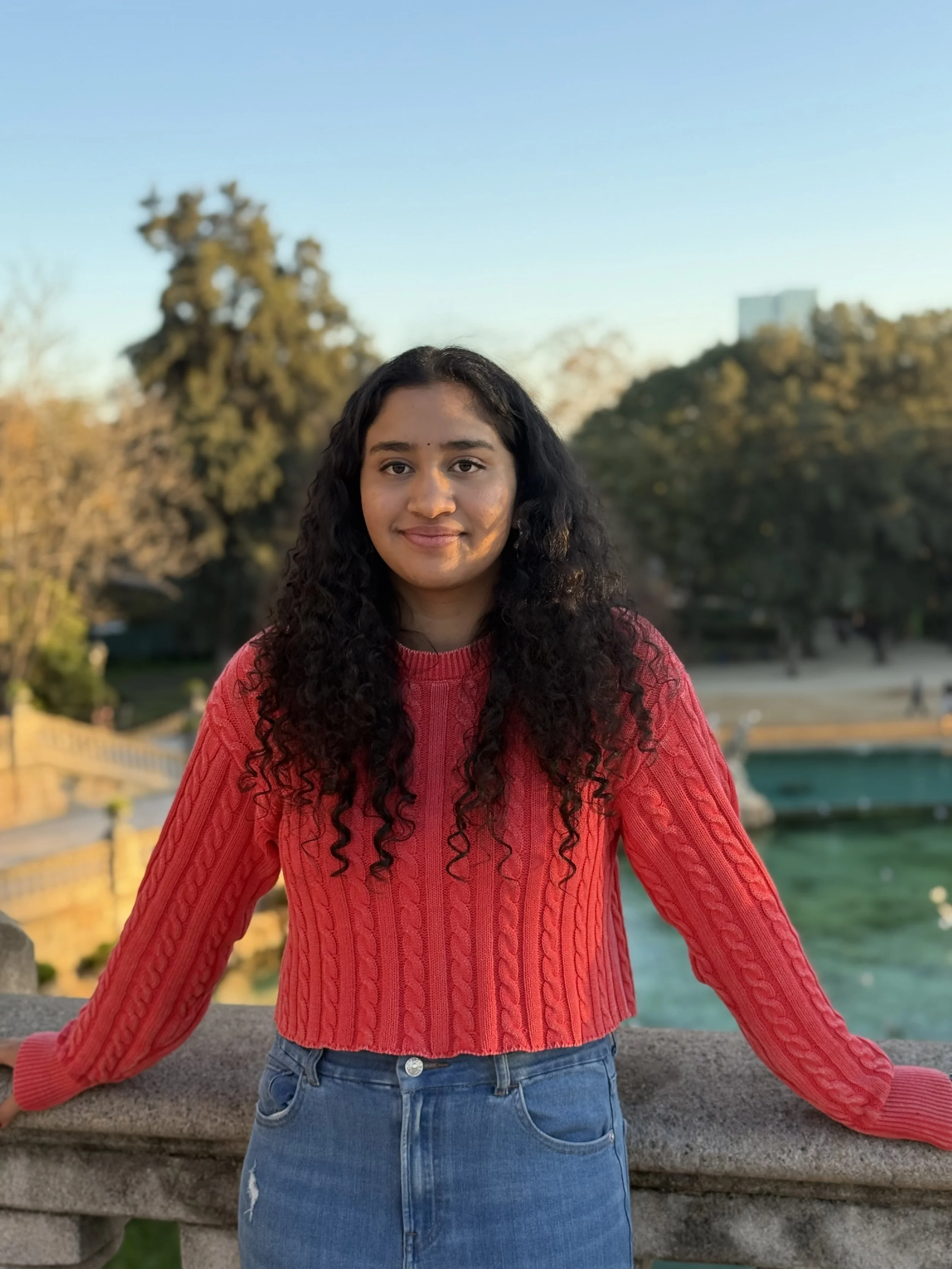 A young woman with long, curly dark hair and a warm smile, wearing a pink cable-knit sweater and blue jeans, standing outdoors by a stone railing near a body of water during sunset.