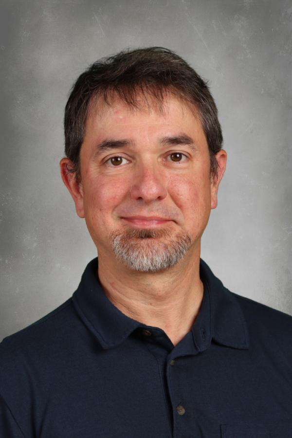 A middle-aged man with short brown hair, a goatee, and light skin, wearing a dark blue collared shirt, standing against a neutral background.