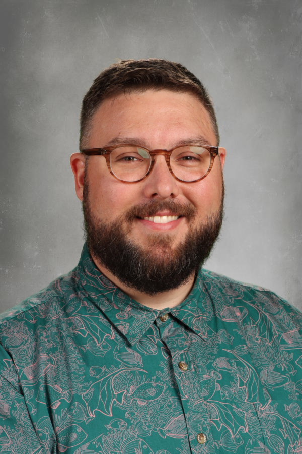 A man with short brown hair, glasses, and a beard smiling in front of a gray background. He is wearing a teal shirt with an aquatic-themed pattern including fish and marine life.