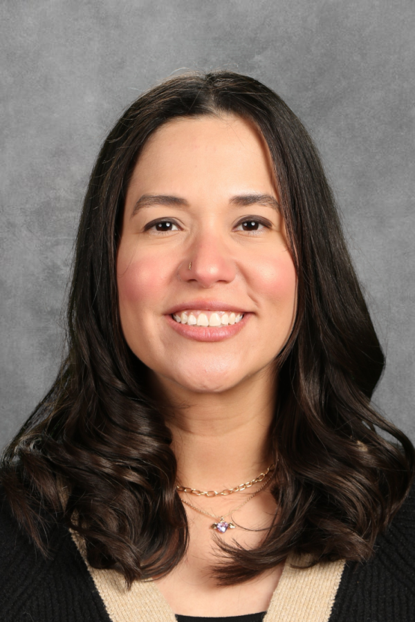 A woman with long dark hair, smiling, wearing a black top with beige accents, and layered necklaces, against a gray background.