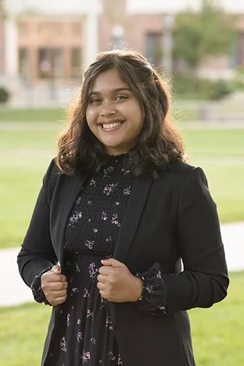 Smiling woman with shoulder-length hair, wearing a black blazer and a floral dress, standing outdoors on a grassy area with blurred buildings in the background.