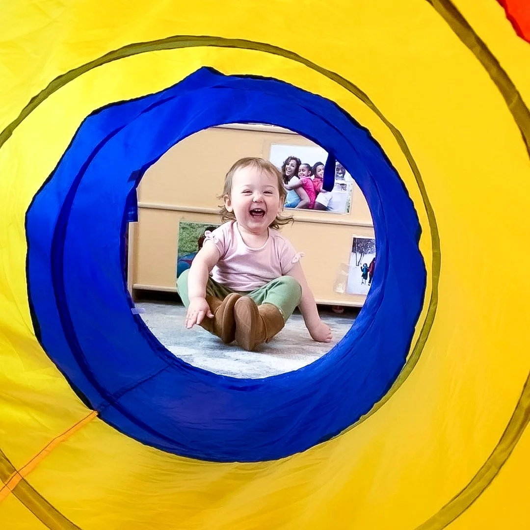 Child laughing and sitting inside a colorful play tunnel.