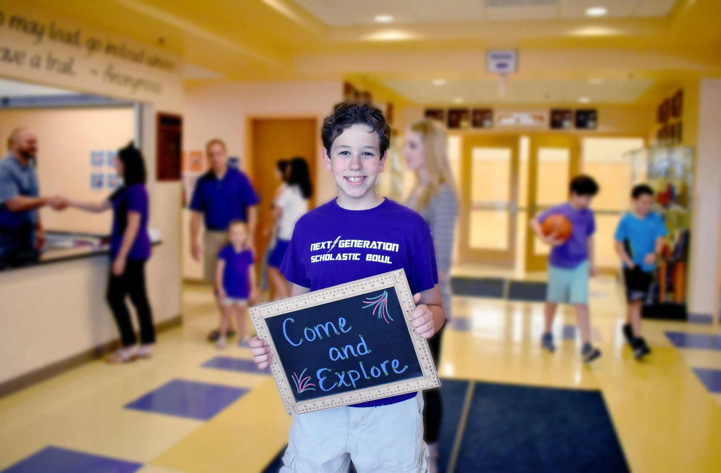 A young boy smiling and holding a sign that says 'Come and Explore' in a school lobby, with other children and adults in the background.