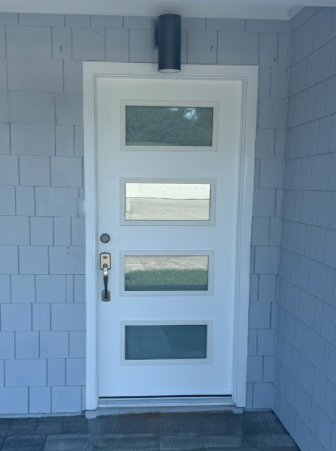 White exterior door with four rectangular glass panels, black handle and lock, and white frame, set in a light blue shingled wall with gray trim and a black cylindrical porch light above.