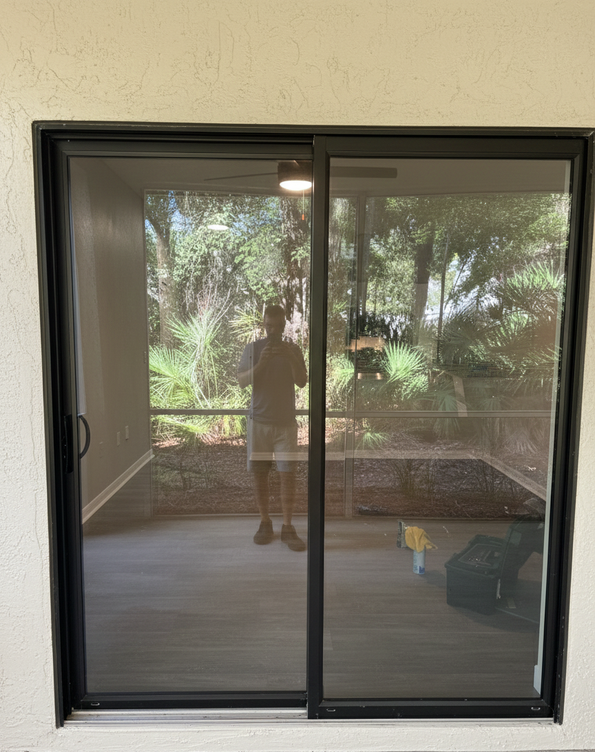 A sliding glass door with black frame, showing a reflection of a person taking a photo, standing on a wooden deck with greenery outside.