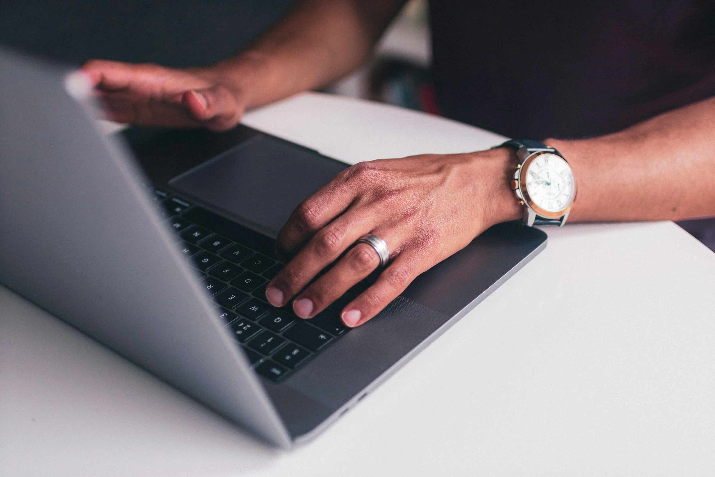 Person typing on a laptop keyboard, wearing a watch and a silver ring on the left hand.