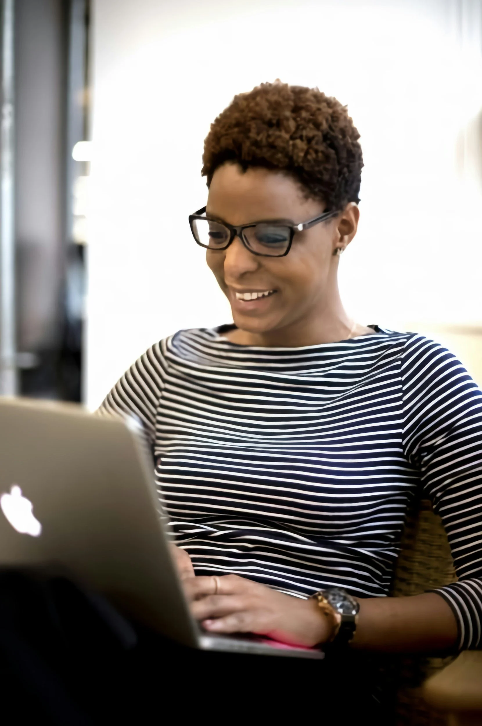 A woman with short curly hair, wearing glasses and a black-and-white striped shirt, smiling while working on a laptop.