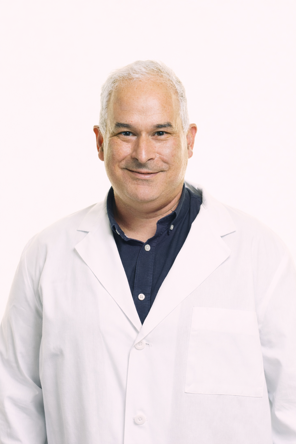 A middle-aged man with short gray hair smiling, wearing a white lab coat over a navy blue button-up shirt, standing against a plain white background.