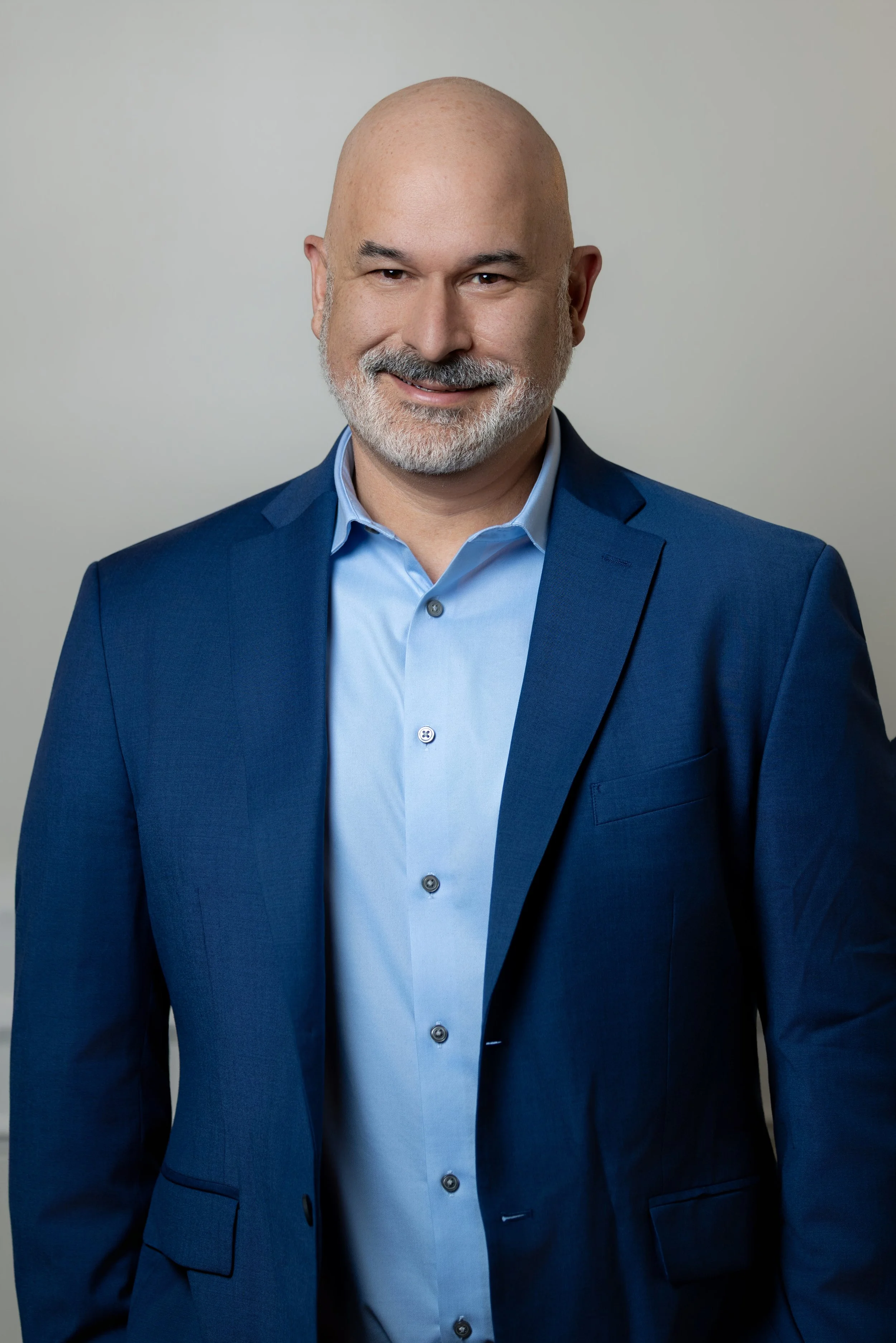 A smiling middle-aged man with a beard and bald head, dressed in a blue suit and light blue shirt, standing against a plain light gray background.