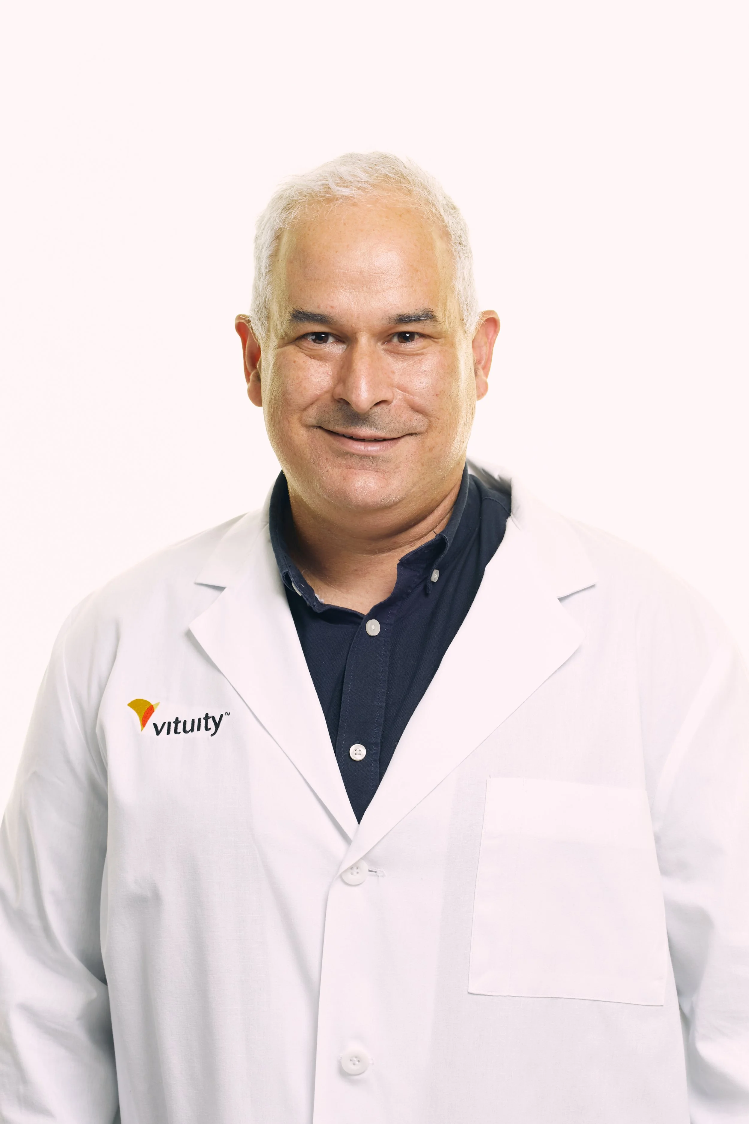 A male doctor with short gray hair smiling at the camera, wearing a white lab coat with 'vitutiy' logo, and a dark blue shirt underneath, standing against a plain white background.