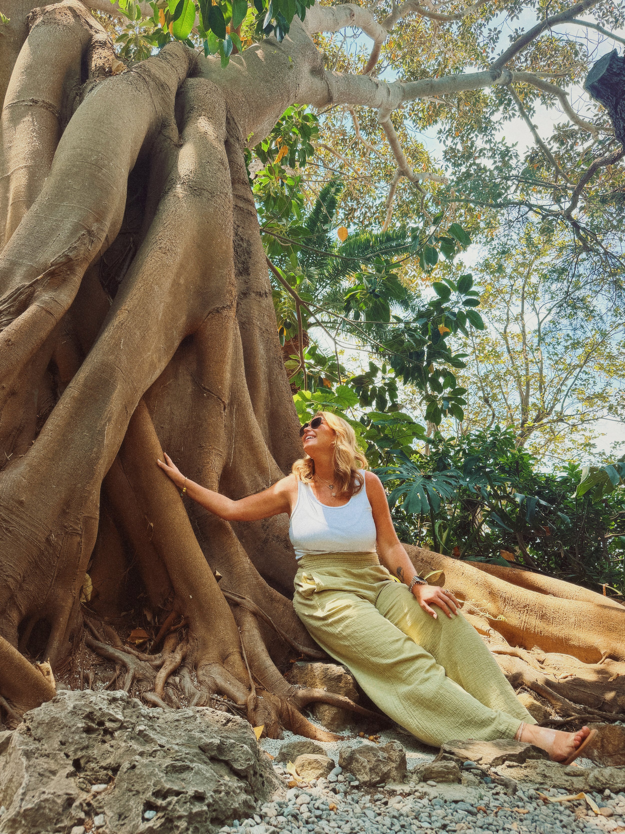 A woman with blonde hair, sunglasses, a white tank top, and beige pants is sitting next to a large, old tree with extensive roots. She is outdoors in a lush, green environment with a clear sky above, touching the tree trunk and smiling.
