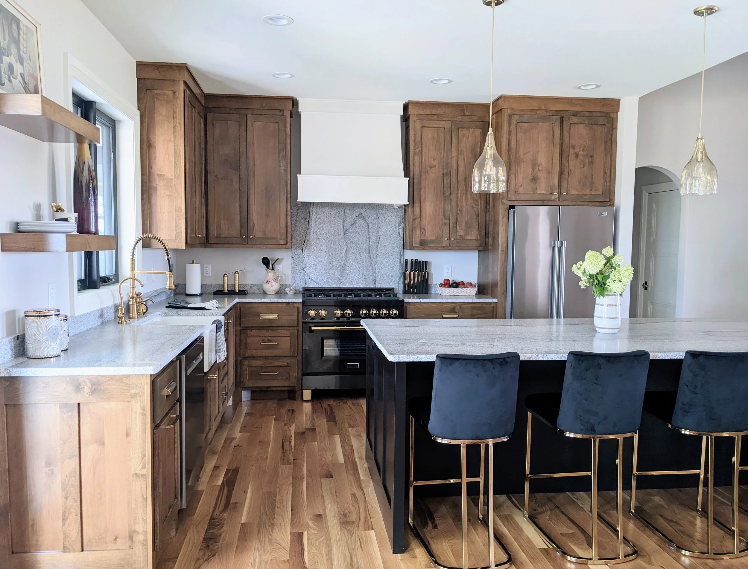 A modern kitchen with wooden cabinets, a white island, a black stove, a stainless steel refrigerator, pendant lights, and a vase with flowers on the island.