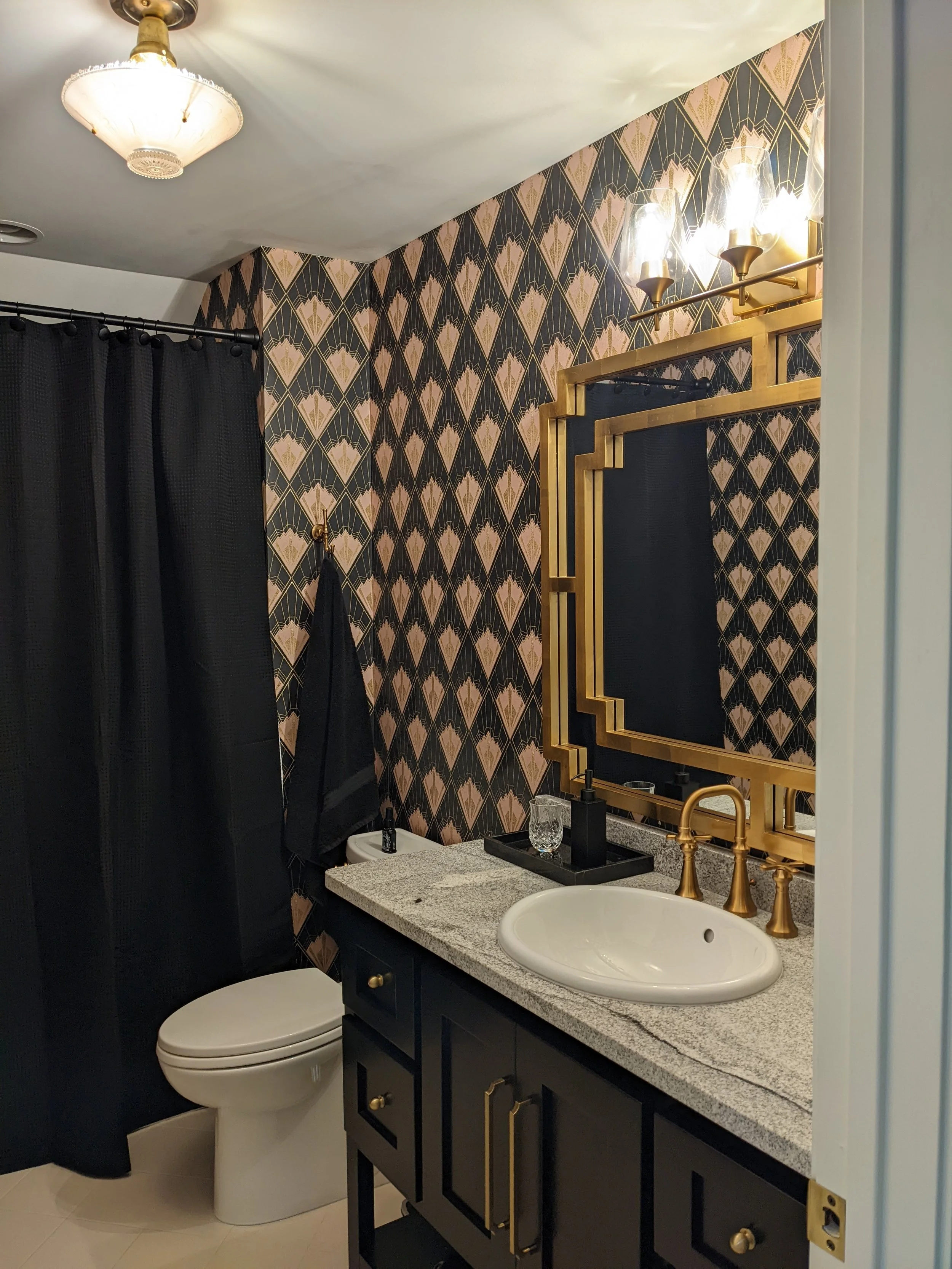 A bathroom with black wallpaper featuring rose gold geometric patterns, a granite countertop with a white sink, a gold-framed mirror, black and gold fixtures, a black vanity.