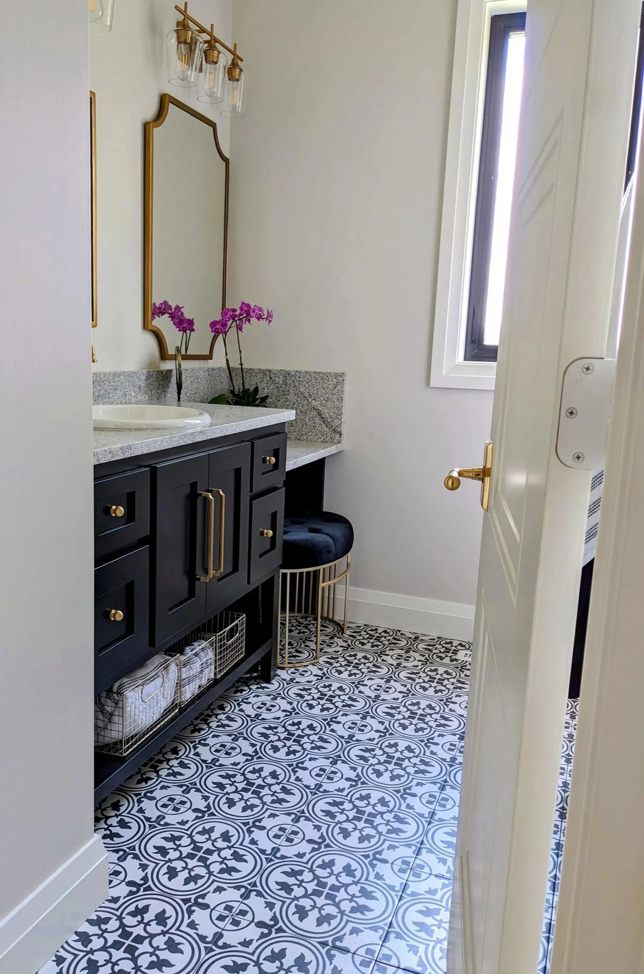 Decorative bathroom with patterned black and white floor tiles, a black vanity with gold accents, a gray granite countertop, and a mirror with a gold frame. There are purple orchids on the counter, a black stool with gold legs, and a window with white framing letting in natural light.