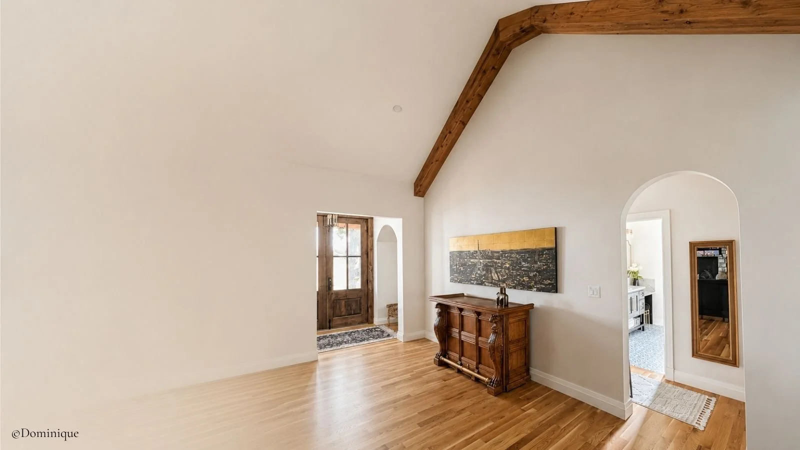 Empty living room with hardwood floors, white walls, a high ceiling with wooden beams, and two doorways leading to other rooms. There is a vintage wooden sideboard against the wall and a long abstract painting above it.