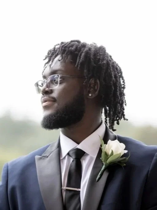 A young man with dreadlocks, glasses, and a beard wearing a blue suit, white shirt, gray tie, and a white boutonniere, looking to the side outdoors.