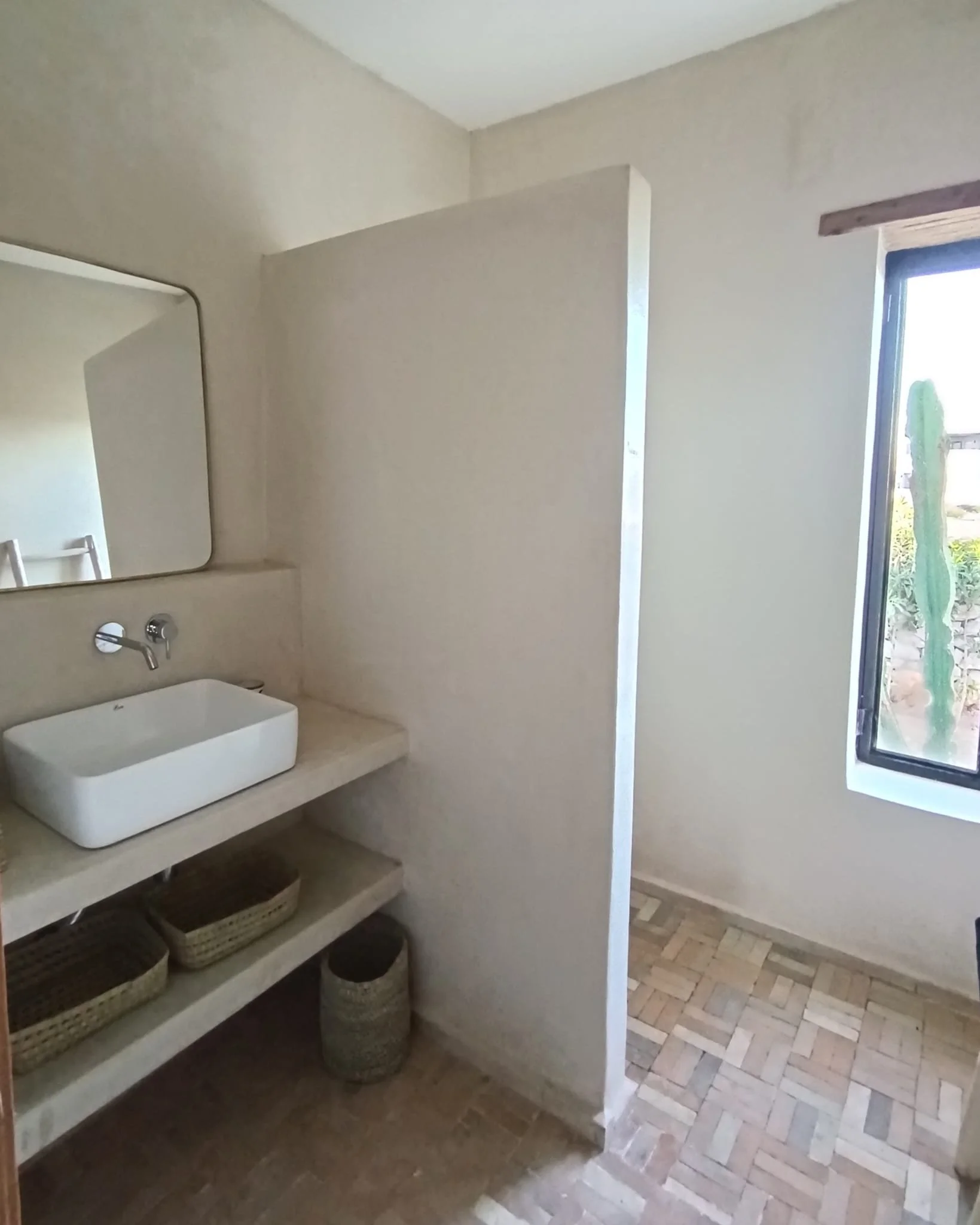 Intérieur d'une salle de bain avec un lavabo carré posé sur une étagère en béton, un miroir rectangulaire au mur, une fenêtre avec vue sur un cactus, et un sol en carreaux de bois.