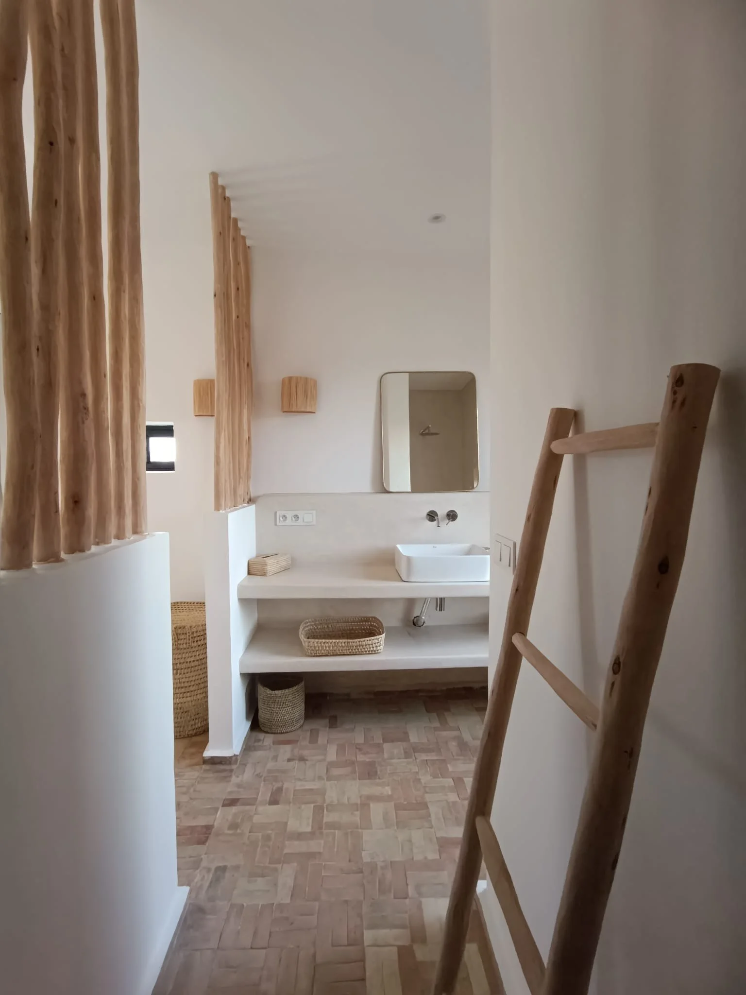 Salle de bains minimaliste avec lavabo carré, miroir rectangulaire, étagère avec paniers en osier, décor en bois naturel, parquet en bois clair, murs blancs, éclairage doux.
