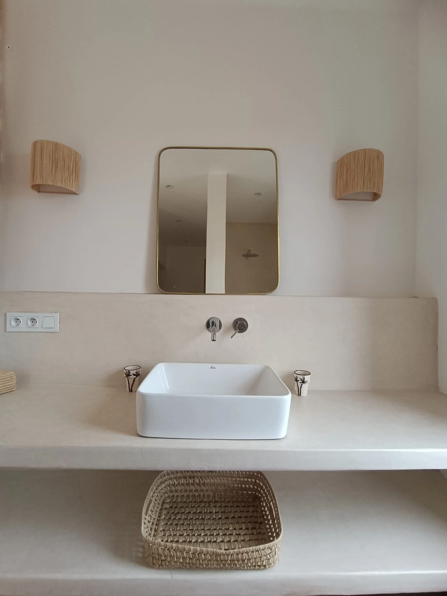 Salle de bain minimaliste avec un lavabo carré blanc, miroir rectangulaire, deux appliques murales en bambou, deux verres décoratifs, prises électriques, et une étagère avec panier en osier dessous.