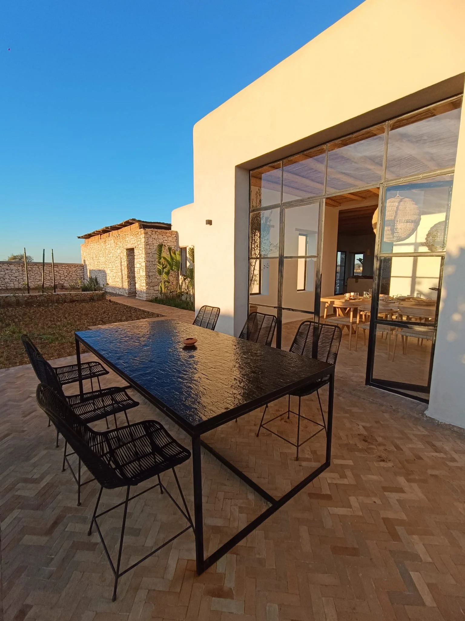 Terrasse extérieure avec une grande table en verre noir et six chaises en métal, vue sur une maison moderne avec grande baie vitrée et une ancienne cabane en pierre en arrière-plan, sous un ciel clair au coucher du soleil.