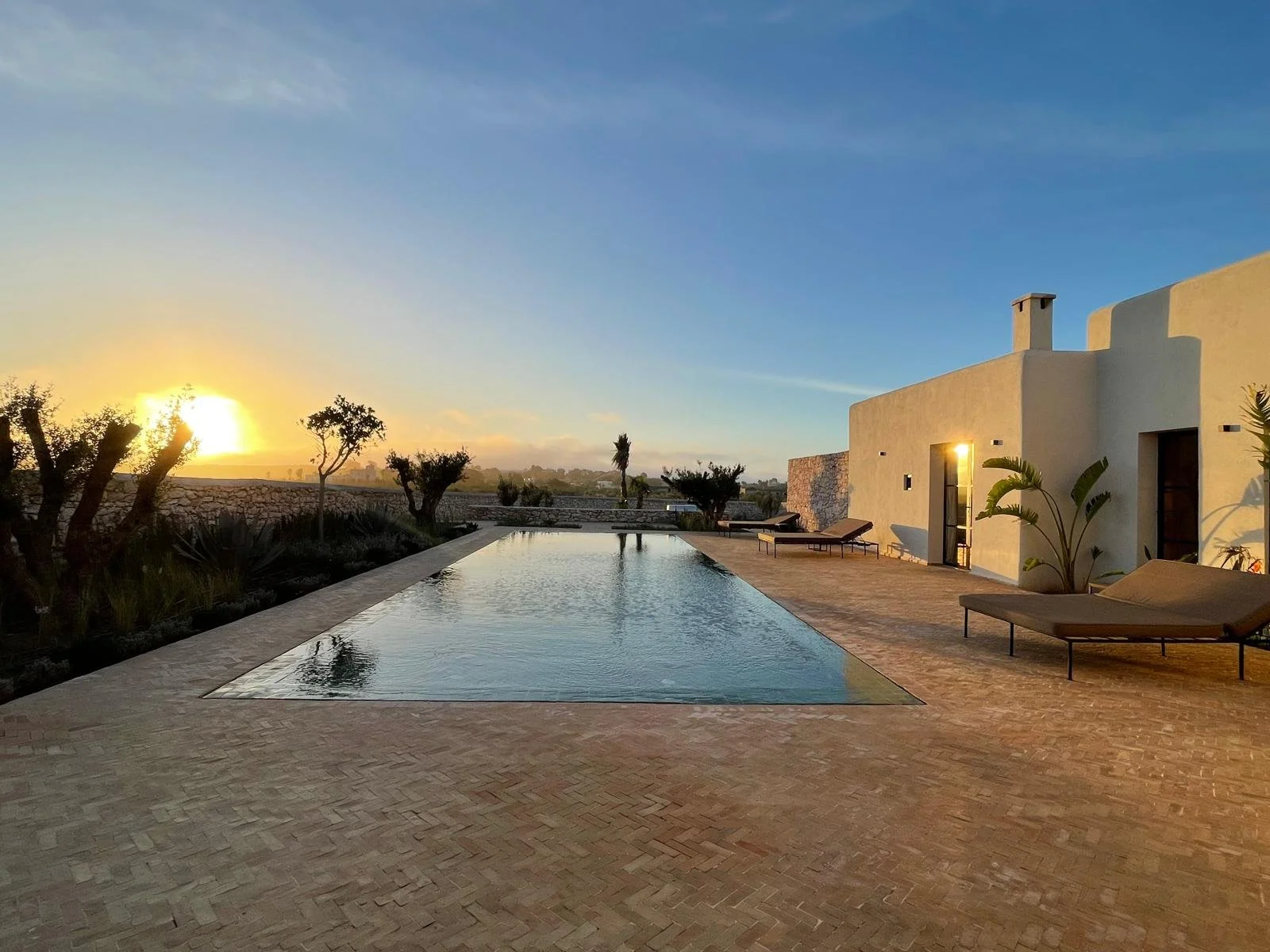 Piscine à côté d'une maison moderne blanche, avec chaises longues, sous un ciel au coucher du soleil, dans un paysage désertique avec des arbres et des plantes tropicales.