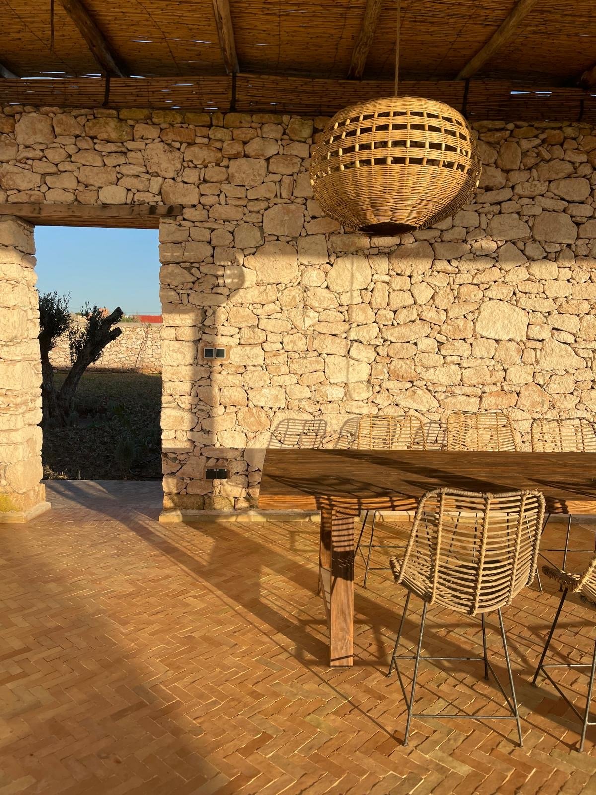 Salle à manger en plein air avec table en bois, chaises en rotin, mur en pierres, lampe en rotin au plafond, éclairée par la lumière du soleil.