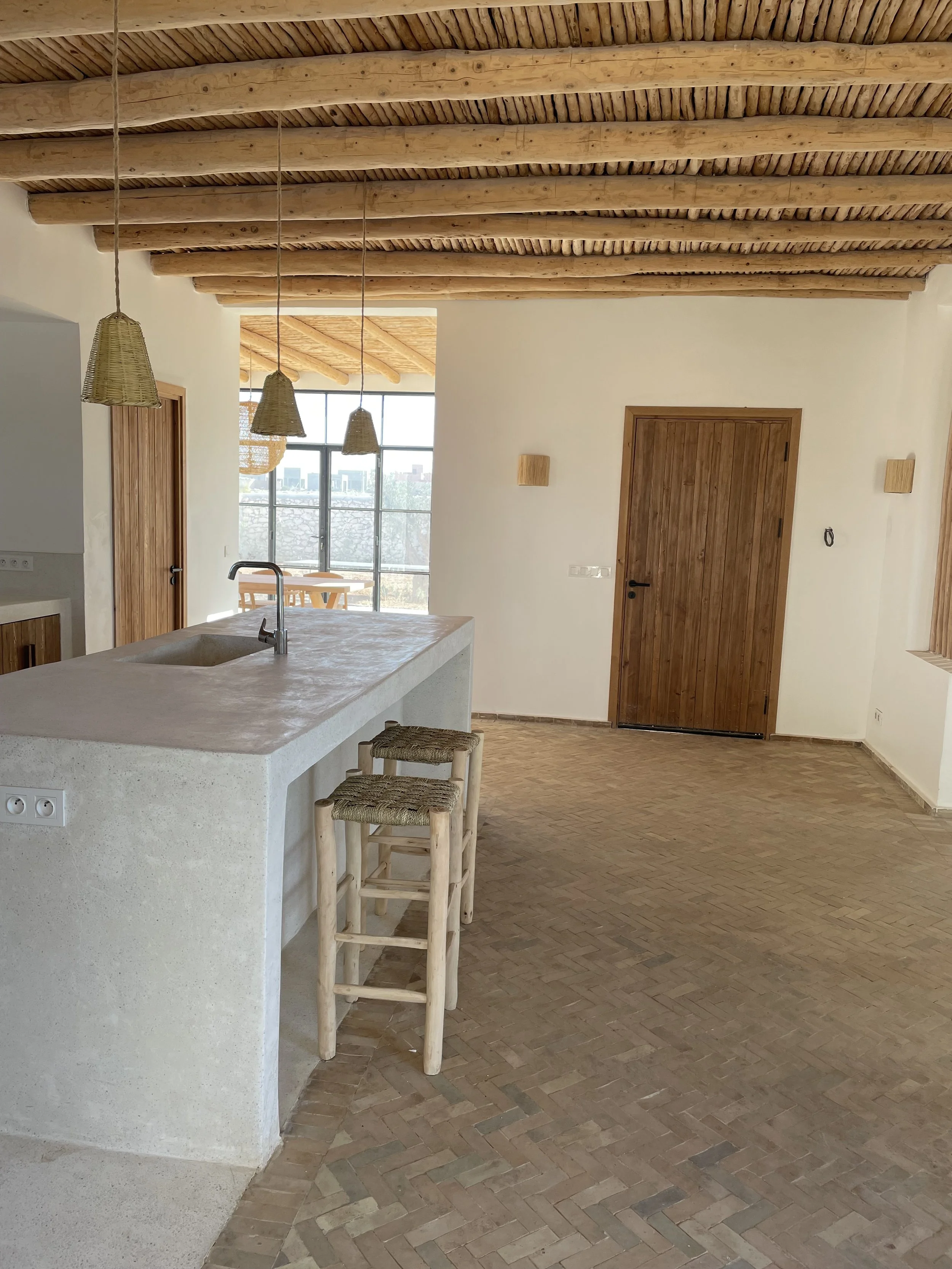 Intérieur d'une cuisine ouverte avec un plan de travail en béton, deux chaises en bois avec des sièges en rotin, un plafond en bois avec des poutres apparentes, un mur blanc avec une porte en bois, une grande fenêtre laissant entrer de la lumière nat