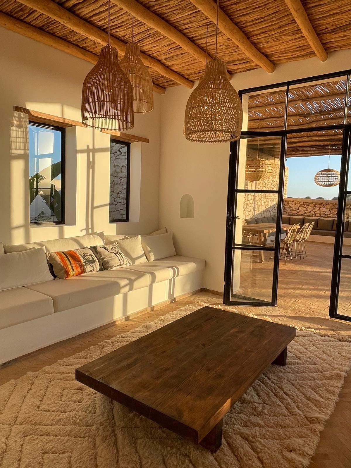Salon intérieur avec un canapé blanc, un tapis beige, une table en bois, des classiques de lumières en rotin suspendues, et une porte-fenêtre donnant sur une terrasse extérieure ensoleillée.