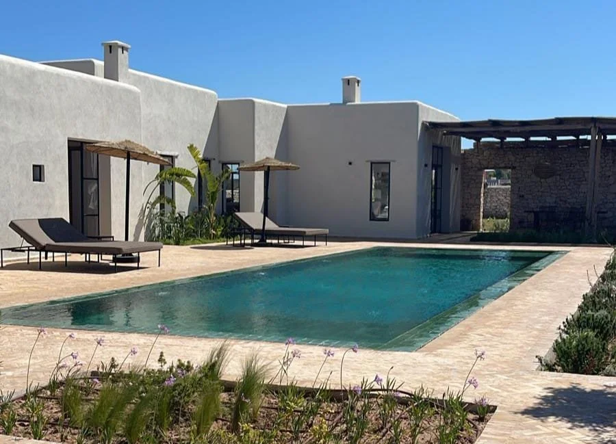 Piscine extérieure dans un espace privé avec transats, plantes et un bâtiment blanc sous un ciel bleu
