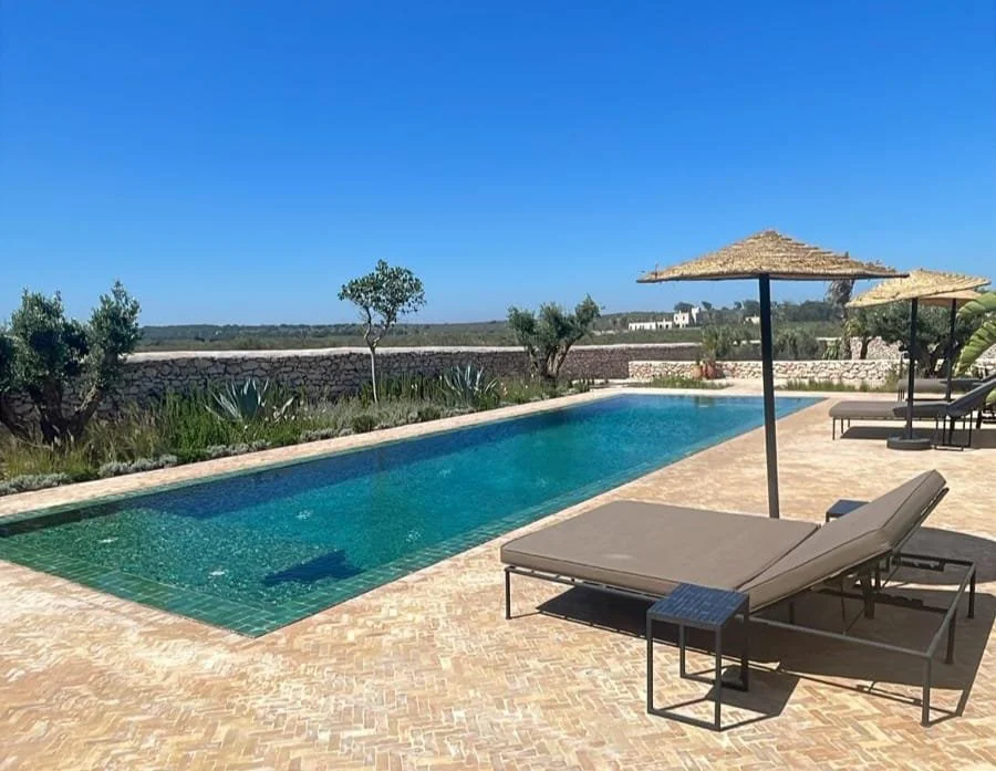 Piscine extérieure avec transats et parasols en paille, vue sur un paysage ouvert et ensoleillé.