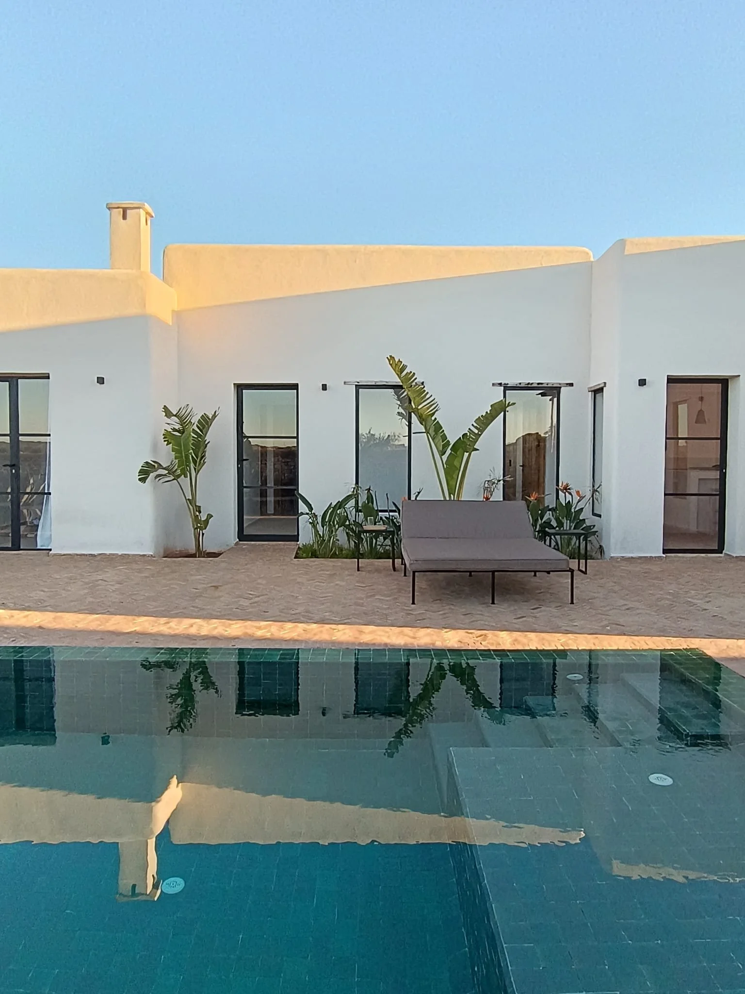 Vue d'une maison blanche moderne avec une piscine à l'avant, des plantes tropicales et un mobilier de terrasse, sous un ciel bleu.