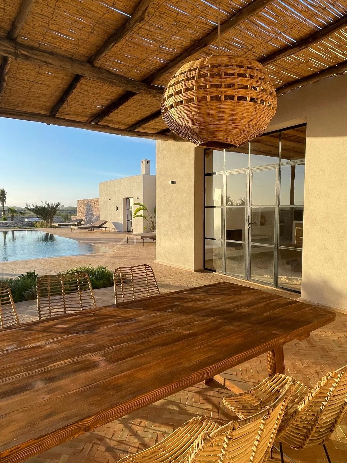 Terrasse avec table en bois, chaises en rotin, vue d'une piscine et maison moderne en plein air lors du coucher du soleil.