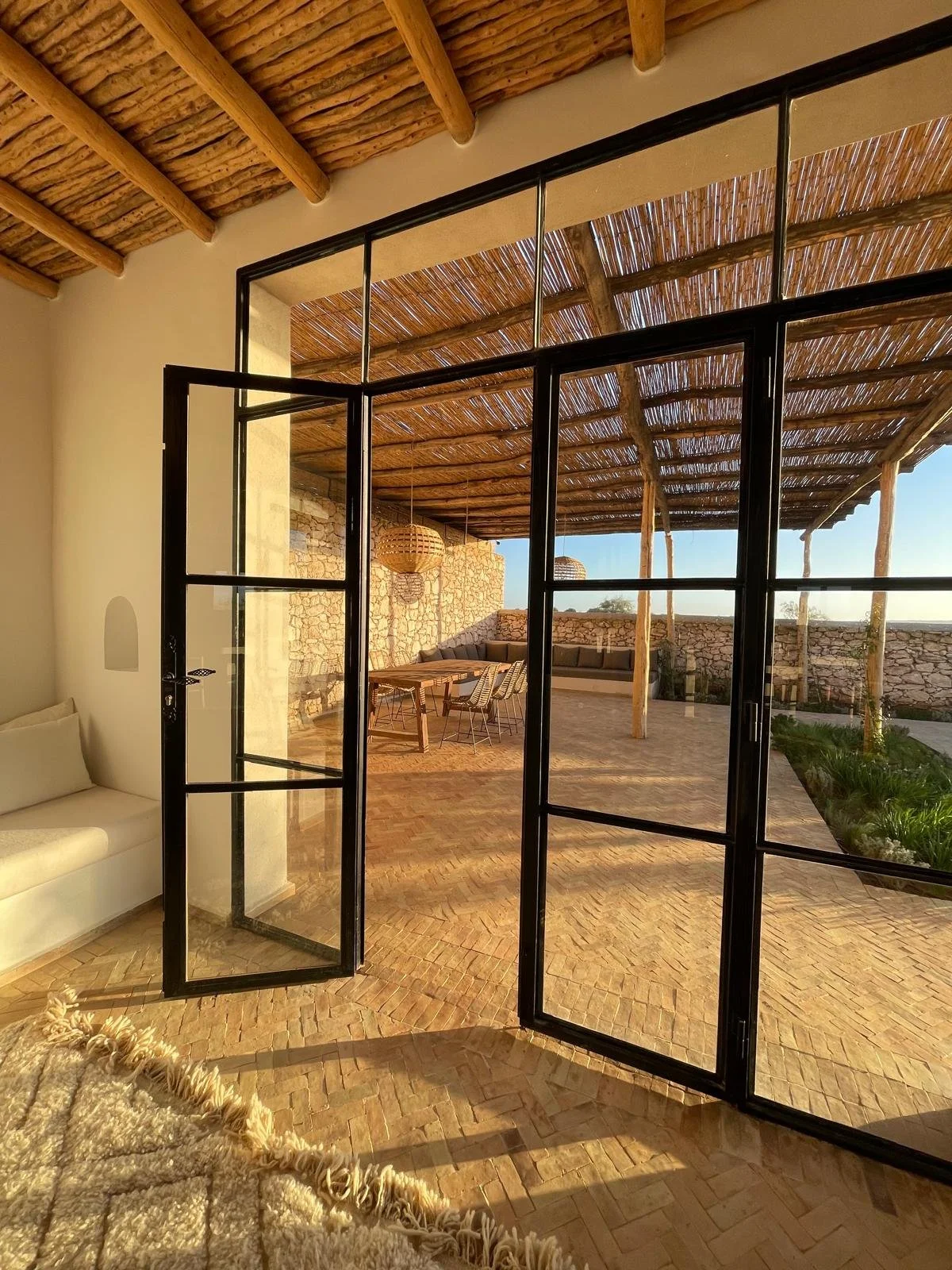 Vue d'une pièce intérieure donnant sur une terrasse extérieure recouverte d'une toiture en matériaux naturels, avec des meubles en bois et un mur en pierre, sous un ciel ensoleillé.