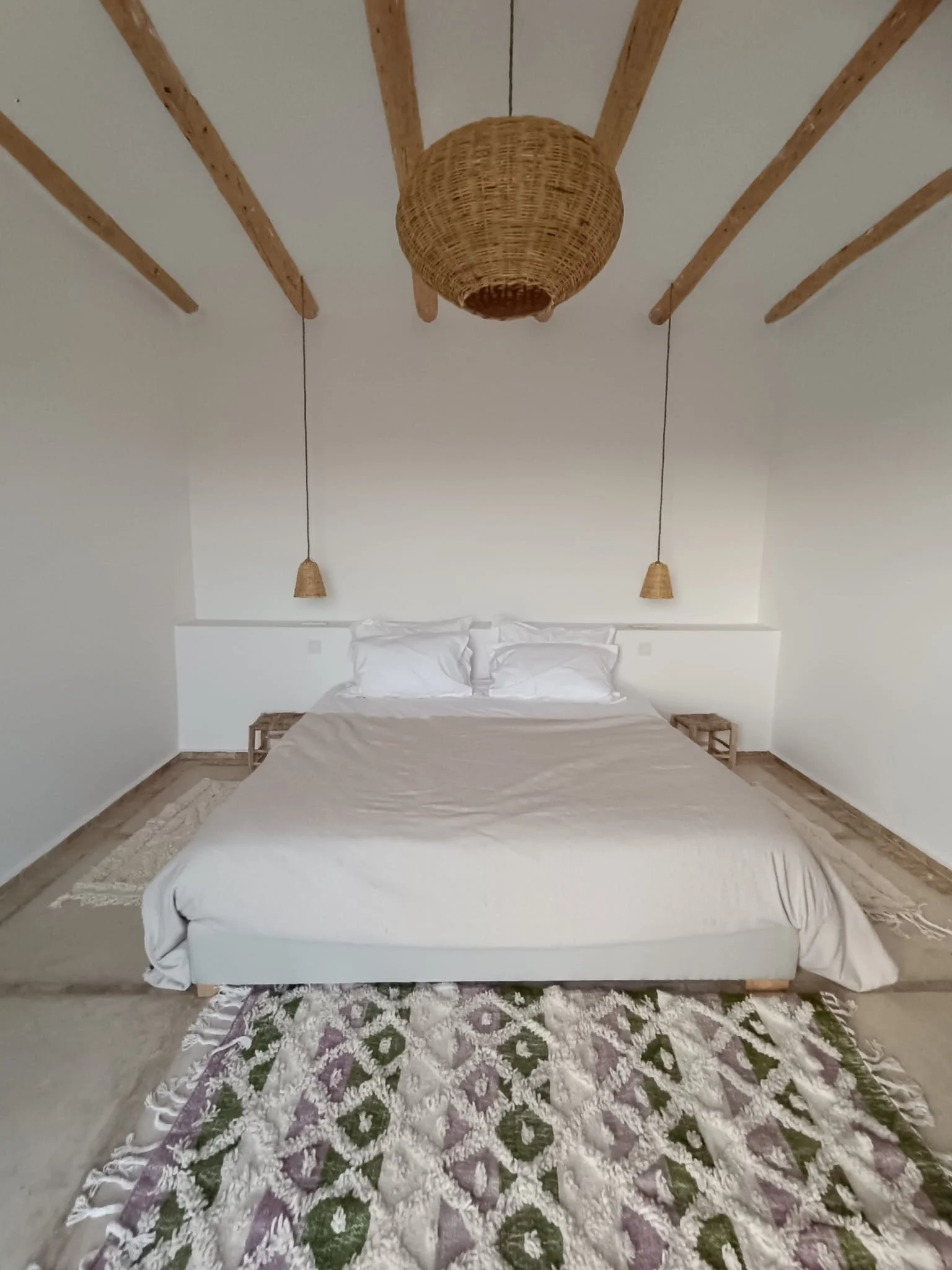 Chambre minimaliste avec lit blanc, deux petites tables en bois, tapis berbère à motifs, suspension en tissu en cimaise avec plafond en bois